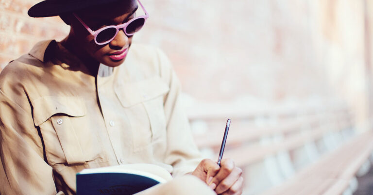 A writer jots an idea down in their notebook, dressed in a monochromatic white outfit, leaning against a white wall.